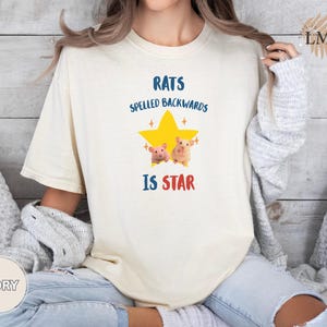 May include: Ivory-colored t-shirt with the text "RATS SPELLED BACKWARDS IS STAR" in blue and red. A yellow star graphic with two cartoon hamsters is centered on the shirt. The shirt is a relaxed fit.