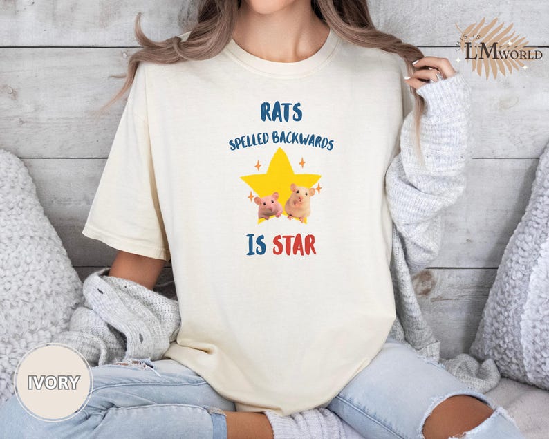 May include: Ivory-colored t-shirt with the text "RATS SPELLED BACKWARDS IS STAR" in blue and red. A yellow star graphic with two cartoon hamsters is centered on the shirt. The shirt is a relaxed fit.