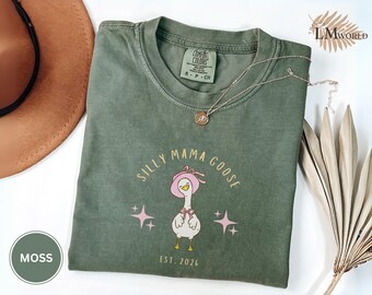 May include: A moss green t-shirt with the words "SILLY MAMA GOOSE" and a cartoon goose wearing a pink hat and bow. The shirt also features the text "EST. 2026" and two pink starbursts. A gold necklace is draped across the shirt.