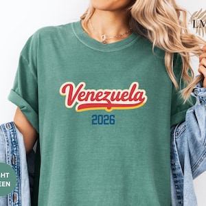 Pu&ograve; includere: T-shirt verde chiaro con la scritta "Venezuela" in corsivo rosso e giallo, e il numero "2026" in blu. La maglietta &egrave; in materiale morbido e ha le maniche arrotolate. La persona indossa una giacca di jeans.