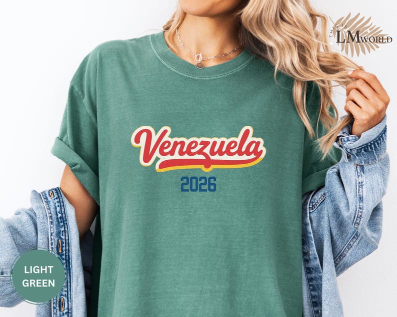Pu&ograve; includere: T-shirt verde chiaro con la scritta "Venezuela" in corsivo rosso e giallo, e il numero "2026" in blu. La maglietta &egrave; in materiale morbido e ha le maniche arrotolate. La persona indossa una giacca di jeans.
