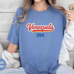 Pu&ograve; includere: Una maglietta blu jeans con la scritta "Venezuela" in rosso e giallo, e l'anno "2026" in blu. La maglietta &egrave; a maniche corte e girocollo.