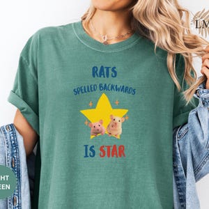 May include: Light green t-shirt with the text "RATS SPELLED BACKWARDS IS STAR" in blue and red. A yellow star with two cartoon rats is in the center. The shirt is a solid color and has rolled-up sleeves.