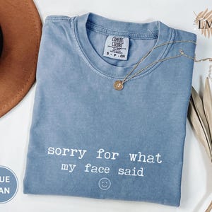 May include: A blue jean Comfort Colors t-shirt with the text "sorry for what my face said" and a smiley face. A gold necklace with a pendant is draped across the shirt. A brown hat and dried plant are in the background.