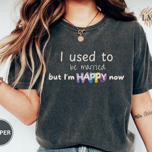 May include: A charcoal gray short-sleeve t-shirt featuring the white text I used to be married but I'm HAPPY now, with the word HAPPY displayed in colorful, patterned block letters. The shirt has a crew neckline and a vintage-style wash.