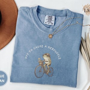 May include: Blue jean Comfort Colors t-shirt with a frog riding a bicycle illustration. The text "OFF TO CAUSE A KERFUFFLE" is arched above the frog. A gold necklace and a brown hat are also in the image.