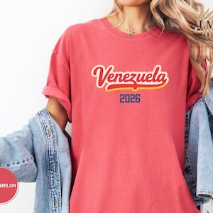 Pu&ograve; includere: Una maglietta rossa con la scritta "Venezuela" in corsivo con contorno giallo e riempimento rosso. Sotto la parola "Venezuela" c'&egrave; il numero "2026" in blu. La maglietta &egrave; indossata con una giacca di jeans.