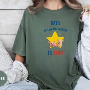 May include: Moss green t-shirt with the text "RATS SPELLED BACKWARDS IS STAR" in blue and red. The design includes a yellow star with two cartoon rats. The shirt is a relaxed fit.