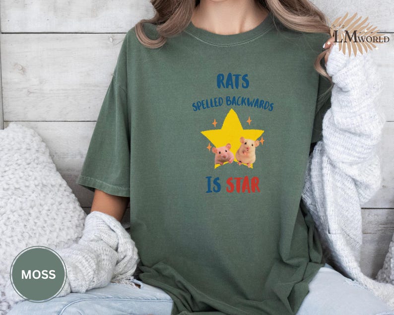 May include: Moss green t-shirt with the text "RATS SPELLED BACKWARDS IS STAR" in blue and red. The design includes a yellow star with two cartoon rats. The shirt is a relaxed fit.