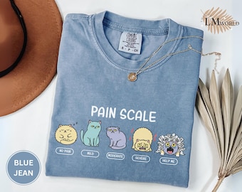 May include: Blue jean colored t-shirt with a "Pain Scale" graphic featuring cartoon cats. The shirt has a crew neck and short sleeves. The graphic includes the text "No Pain", "Mild", "Moderate", "Severe", and "Help Me". A gold necklace is draped across the shirt.