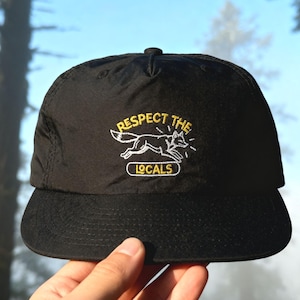 May include: A black baseball cap featuring an embroidered graphic of a running fox in white thread. Above the fox, the text reads RESPECT THE in yellow, and below the fox, the word LOCALS is embroidered in yellow inside a white outlined box.