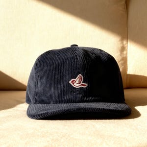 May include: A dark navy blue corduroy baseball cap featuring a small, embroidered patch on the front panel. The patch depicts a stylized bird in white and red thread, shown in profile with wings extended as if in flight against the textured fabric.