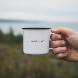 May include: White enamel camping mug with black rim and handle, speckled with black dots. The mug features the text "woods > city" in a minimalist font. The mug is held by a hand, with a blurred outdoor background.