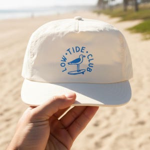 May include: An off-white baseball cap featuring a blue circular graphic on the front panel. The graphic depicts a seagull standing on a surfboard with the text LOW TIDE CLUB printed in a circular arrangement around the central bird illustration.