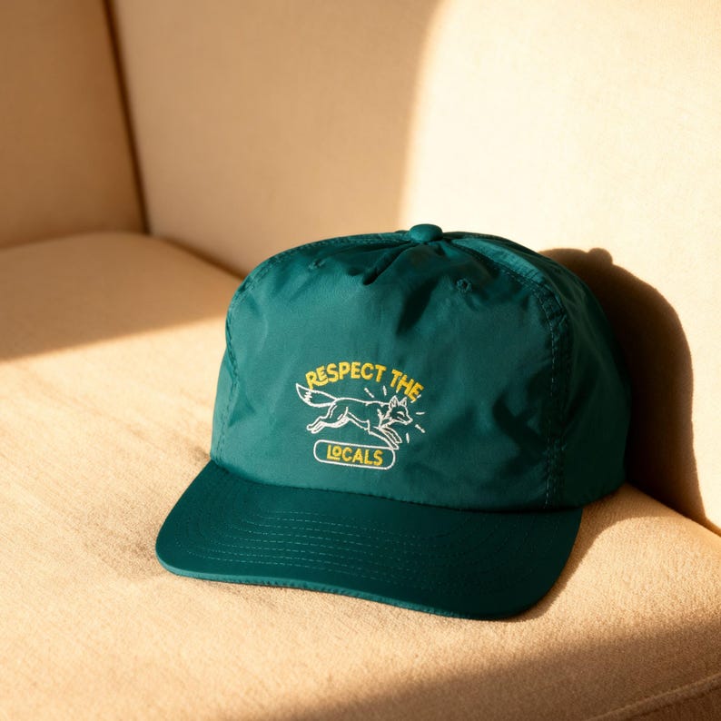 May include: A dark green baseball cap featuring an embroidered graphic of a running fox in white thread. Above the fox, the text reads RESPECT THE in yellow, and below the fox, the word LOCALS is enclosed in a yellow rectangular outline.