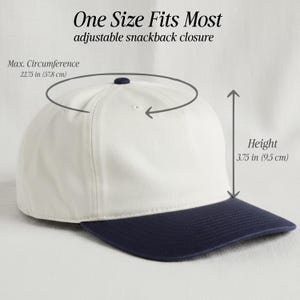 May include: A white baseball cap with a dark blue brim and an adjustable snapback closure. The cap features a maximum circumference of 22.75 inches and a height of 3.75 inches. Text on the image reads One Size Fits Most, adjustable snapback closure.