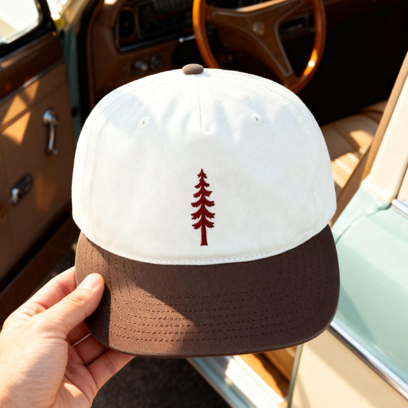 May include: A baseball cap featuring a white crown and a brown brim. The front of the cap displays a small, embroidered dark red pine tree icon centered on the white fabric. The cap has a brown button at the top center and visible stitching.