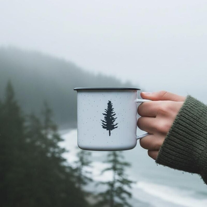 May include: White enamel mug with a black rim and a black pine tree design. The mug has a speckled pattern and is held by a person wearing a green sweater. The background is a misty outdoor scene.
