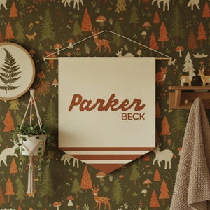 May include: A cream-colored fabric wall banner featuring the name Parker BECK in brown script and block lettering, with two horizontal brown stripes at the bottom. The banner hangs from a wooden dowel with a white string against a forest wallpaper.