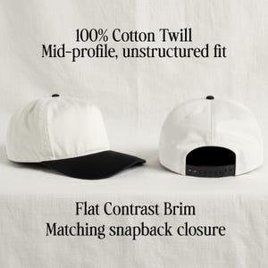May include: A white baseball cap with a black flat brim and a black snapback closure. The cap is made of 100% cotton twill and features a mid-profile, unstructured fit. The text 100% Cotton Twill, Mid-profile, unstructured fit, Flat Contrast Brim, Matching snapback closure is visible.