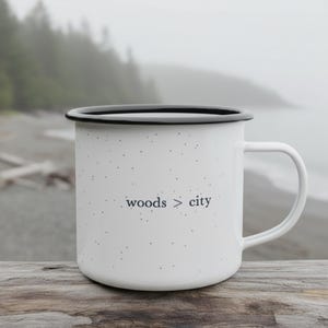 May include: White enamel mug with a black rim and handle. The mug has a speckled pattern and the words "woods > city" printed in black. The mug is sitting on a wooden surface.