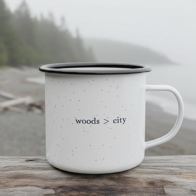May include: White enamel mug with a black rim and handle. The mug has a speckled pattern and the words "woods > city" printed in black. The mug is sitting on a wooden surface.