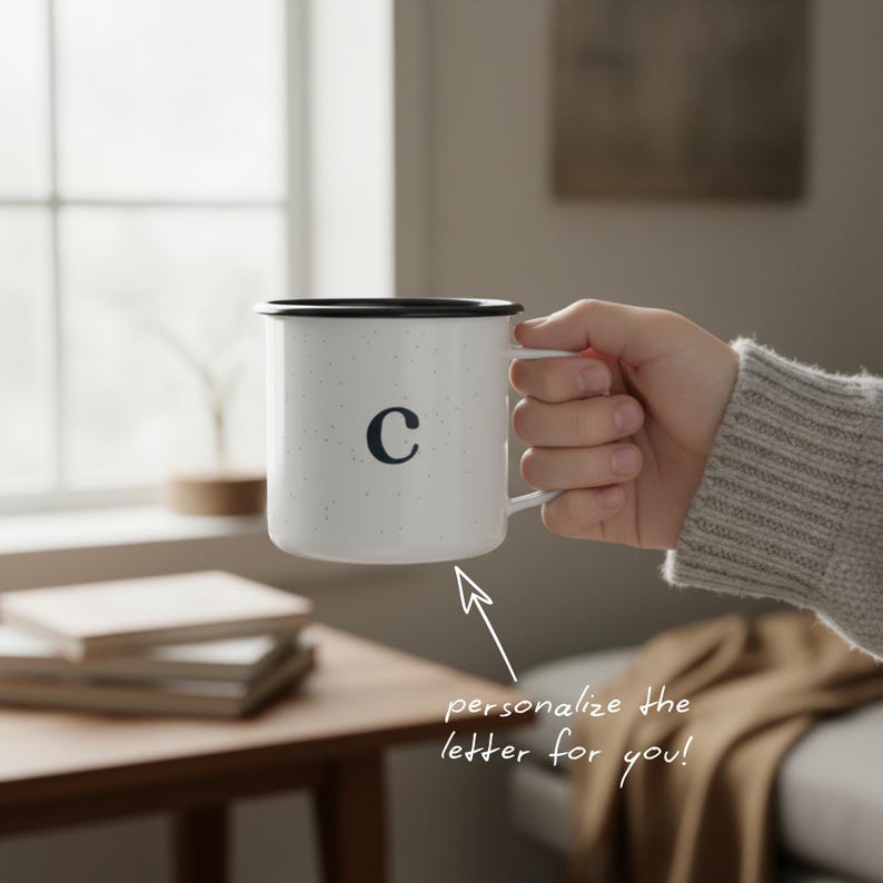 May include: White speckled enamel mug with a black rim and handle, featuring the letter "C" in black. The mug is held in a hand. The text "personalize the letter for you!" is below the mug. A stack of books and a wooden table are in the background.