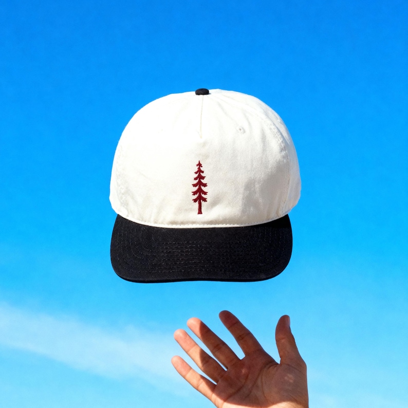 May include: A baseball cap with a white crown and a black brim. The front of the crown features a small, embroidered dark red pine tree design. The cap has a black button at the top center where the panels meet, providing a clean, structured look.