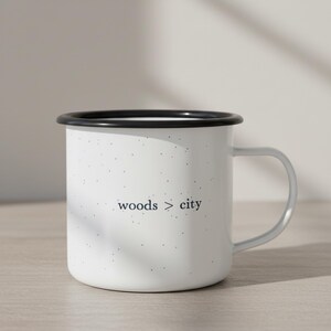 May include: White enamel mug with a black rim and a gray handle. The mug has a speckled pattern and the words "woods > city" printed in a dark gray font. The mug is designed for hot beverages.