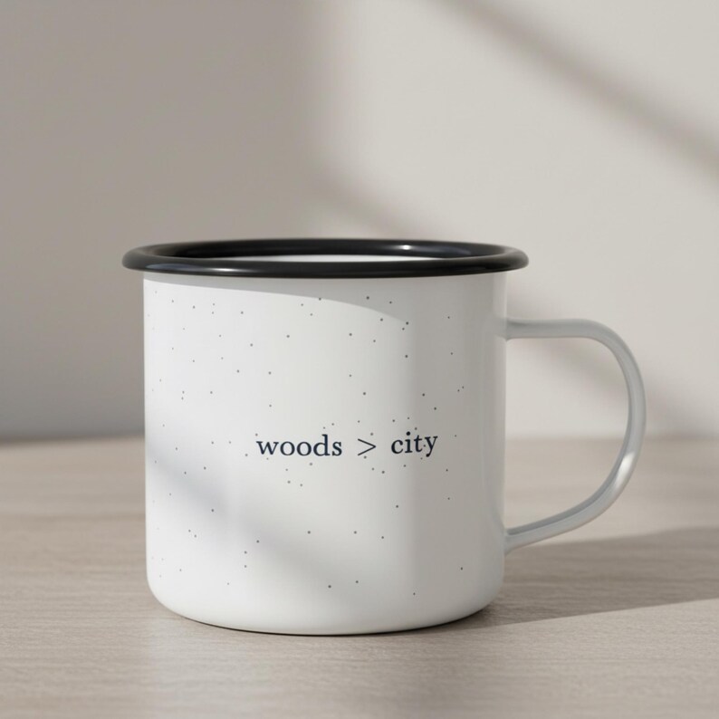 May include: White enamel mug with a black rim and a gray handle. The mug has a speckled pattern and the words "woods > city" printed in a dark gray font. The mug is designed for hot beverages.