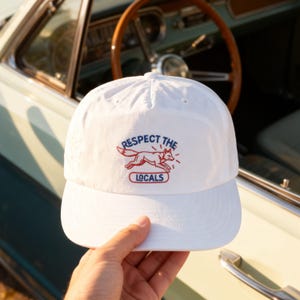 May include: A white baseball cap featuring an embroidered graphic of a red fox in motion. Above the fox, the text reads RESPECT THE in blue, and below the fox, the word LOCALS is enclosed in a red oval outline. The cap has a structured crown.