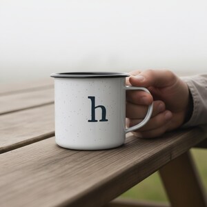 May include: White enamel mug with a black rim and handle, featuring the letter "h" in navy blue. The mug has a speckled design and is held by a person. The mug is sitting on a wooden picnic table.