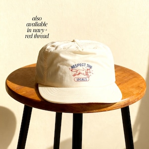 May include: An off-white baseball cap featuring an embroidered design of a red fox in motion. Above the fox, the text reads RESPECT THE in blue, and below the fox, the word LOCALS is enclosed in a blue rectangular outline with red thread.