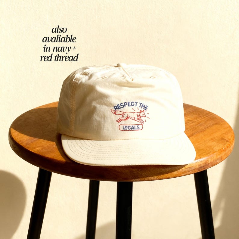 May include: An off-white baseball cap featuring an embroidered design of a red fox in motion. Above the fox, the text reads RESPECT THE in blue, and below the fox, the word LOCALS is enclosed in a blue rectangular outline with red thread.