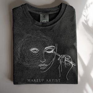 Pode incluir: Uma camiseta cinza escura com um design gráfico branco de um rosto de mulher com um olho cercado por um design de sol. A mulher está aplicando maquiagem com um pincel. O texto "MAKEUP ARTIST" está impresso abaixo do gráfico.
