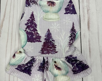 Puede incluir: Un vestido para perro de color morado y blanco con un dobladillo con volantes. El patrón presenta muñecos de nieve, pinos y copos de nieve.