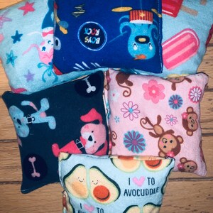 May include: Six colorful fabric squares with different patterns. The patterns include a blue square with a cartoon character playing drums, a pink square with monkeys and flowers, a blue square with avocados and the text "I heart to avo cuddle", a black square with a dog and bone, a blue square with stars and a pink bunny, and a pink square with popsicles and a blue background.