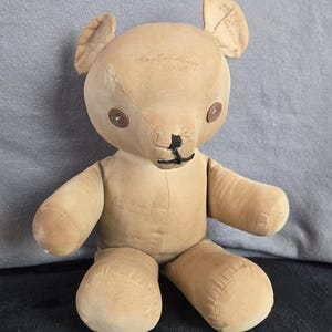 May include: A vintage tan teddy bear with button eyes and a stitched nose. The bear has a classic design with jointed limbs and a soft, plush texture. The bear has text on its head and ear.