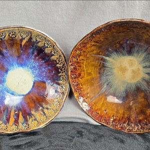 May include: Two ceramic bowls with a glossy finish. The bowls have a circular shape with a central design in shades of blue, purple, and gold. The rims are decorated with a floral pattern. The bowls are approximately 6 inches in diameter.
