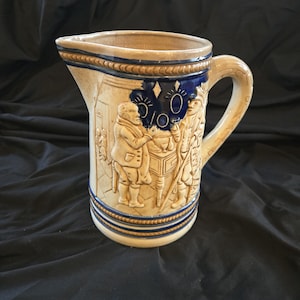 May include: A beige ceramic pitcher with a detailed relief design featuring figures in period clothing. The pitcher has a blue band near the top and bottom, and a handle on the side. The overall style suggests a vintage or antique piece.