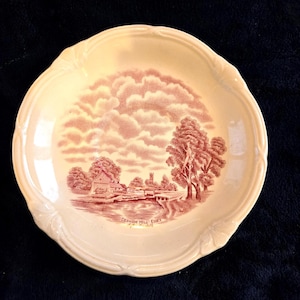 May include: Cream-colored ceramic plate with a scalloped edge and a red transferware design. The design depicts a rural landscape with buildings, trees, and a body of water. The text "Dedham Mill Essex" is printed at the bottom.