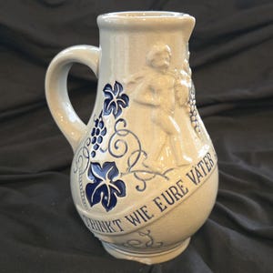 May include: A stoneware pitcher with a handle, featuring a relief design of a man, grapes, and vines. The pitcher is beige with blue accents and the text "TRINKT WIE EURE VATER" on a banner. The pitcher is approximately 9 inches tall.