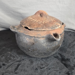 May include: A rustic, handmade ceramic pot with a lid. The pot is a dark gray color with a brown lid and handle. The lid has a decorative pattern. The pot has a spout on the side.