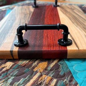 May include: A rectangular serving tray constructed from multi-coloured wood planks featuring distinct grain patterns in shades of light tan, dark brown, and deep red. The tray is equipped with two black metal pipe-style handles on top.