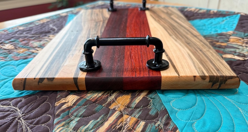 May include: A rectangular serving tray constructed from multi-coloured wood planks featuring distinct grain patterns in shades of light tan, dark brown, and deep red. The tray is equipped with two black metal pipe-style handles on top.