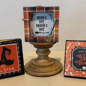 May include: Three square Halloween-themed signs. The left sign is black with a witch hat and the text "too CUTE to SPOOK". The center sign features a plaid frame and the text "MOON'S OUT BROOM'S OUT" with a broom graphic. The right sign has a skeleton pattern and the text "SHAKE YOUR BOO THANG".