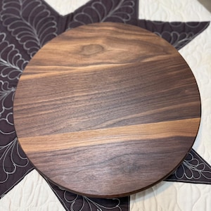 May include: A circular wooden serving board crafted from dark brown walnut wood. The surface displays a natural wood grain pattern with horizontal variations in color, ranging from deep chocolate brown to lighter tan tones across the flat board.