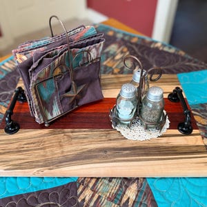 May include: A rectangular wooden serving tray with black metal pipe handles, featuring a metal napkin holder with a star accent and a metal caddy containing two glass salt and pepper shakers on a small white crocheted doily, holding patterned napkins.