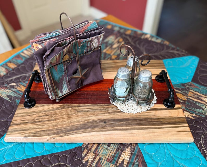 May include: A rectangular wooden serving tray with black metal pipe handles, featuring a metal napkin holder with a star accent and a metal caddy containing two glass salt and pepper shakers on a small white crocheted doily, holding patterned napkins.