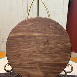 May include: A circular dark brown wooden board with a visible natural grain pattern, displayed on a gold-toned metal stand featuring a pointed finial at the top and curved supports at the base to hold the board in an upright vertical position.